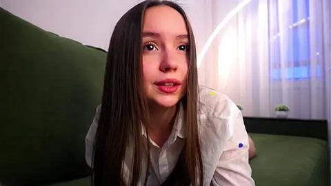 Media: Video of a young woman with straight, long brown hair, fair skin, and a slight smile. She wears a white button-up shirt with colorful polka dots. The background features a green couch and a white curtain with a blue window behind her.