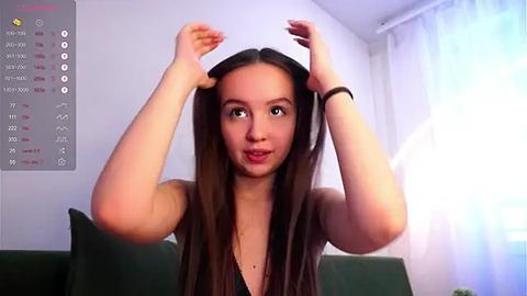 Media: A video of a young woman with long, straight brown hair, wearing a black top, adjusting a black wig in a modern living room with white walls and a window with sheer curtains.