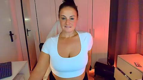 Media: Video of a smiling, light-skinned woman with brown hair tied in a bun, wearing a white crop top, seated in a dimly lit bedroom with white wardrobes and red lighting.