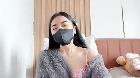 Media: Video of an Asian woman with long black hair, wearing a black face mask, pink lace lingerie, and a gray fuzzy robe, lying on a bed with wooden headboard, beige pillows, and a stuffed teddy bear.