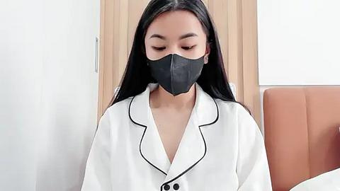 Media: Video of an Asian woman with long black hair wearing a white satin robe with black trim, black mask, and closed eyes in a modern, minimalist bedroom.