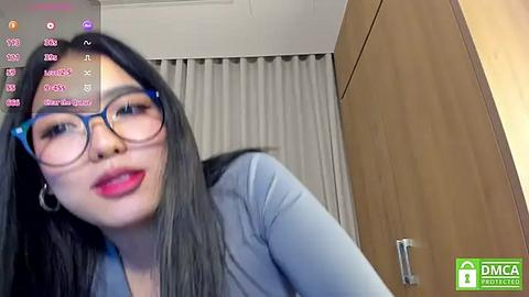 Media: A video of an Asian woman with long black hair, wearing glasses and a blue top, standing in front of a wooden wardrobe and white curtains, with social media icons and text overlay.
