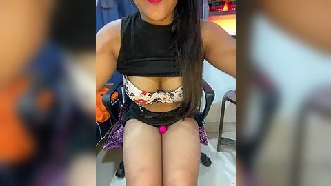 Media: Video of a woman with medium skin tone, wearing a black crop top revealing a patterned bra, sitting on a purple chair. Her long, straight black hair falls over her shoulders. Background includes a blurred, warm-toned room with a sofa and a TV.