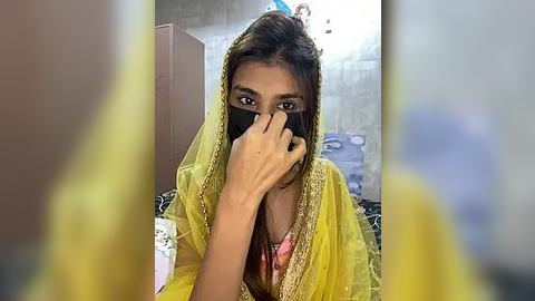 Media: Video of a woman in a yellow sari with a black mask, partially covering her face, sitting on a bed. Background includes a blurred room with a blue car.