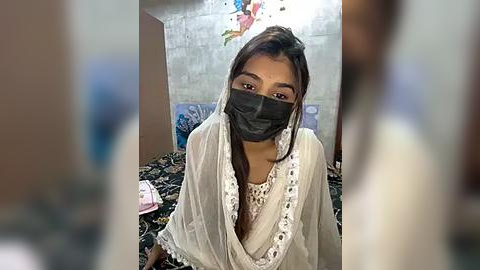 Media: Video of a young South Asian woman in a modest, white embroidered dress, wearing a black face mask, sitting on a bed with floral-patterned sheets, in a simple, dimly lit room.