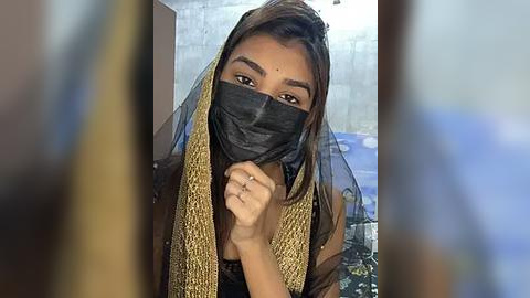 Media: Video of a young South Asian woman with long dark hair, wearing a black mask, gold shawl, and brown top, with blurred background of a concrete wall and blue objects.