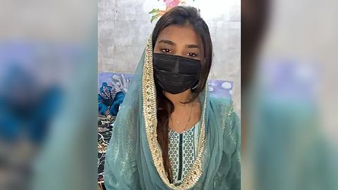 Media: Video of a woman wearing a black face mask, teal shalwar kameez with golden embroidery, and a black hijab, seated indoors against a floral-patterned backdrop.