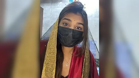 Media: Video of a woman with long dark hair, wearing a black mask and a red sari with gold borders, indoors with a concrete wall background.