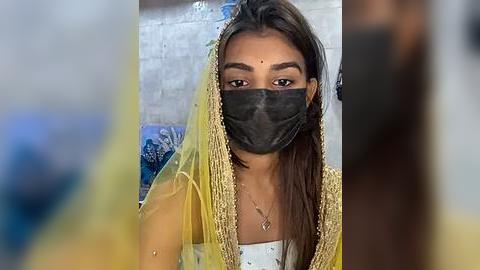 Media: Video of a woman with medium brown skin, long dark hair, and a black face mask, wearing a yellow and gold sari, seated in a dimly lit room with a tiled wall and indistinct background.