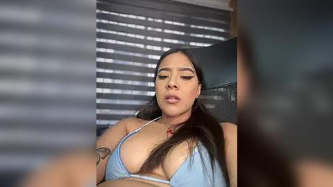 Media: Video of a Latina woman with long dark hair, wearing a light blue bikini, lying on a bed with blinds partially closed in the background.