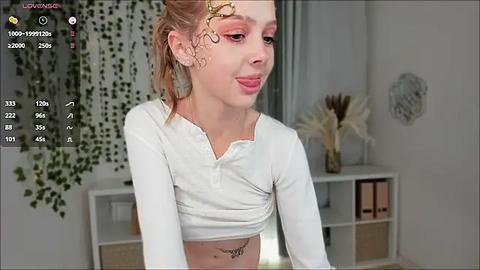 Media: Video of a young, fair-skinned woman with curly blonde hair, wearing a white crop top, standing in a modern living room with green vines and white decor.