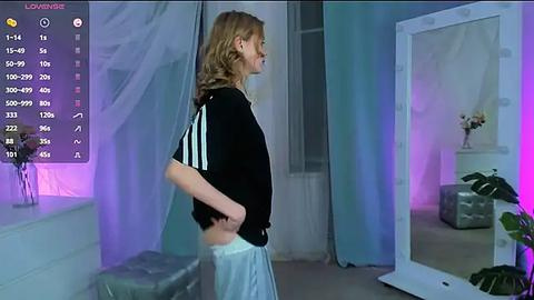 Media: Video of a blonde woman in a black Adidas shirt and white pants, standing in a modern, minimalist room with a mirror, potted plants, and purple lighting.