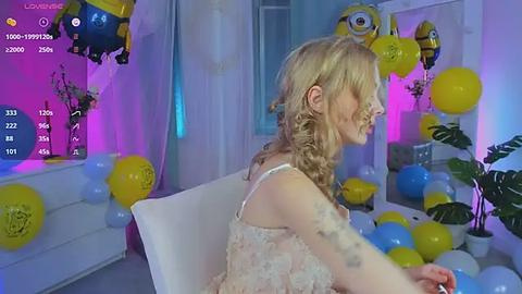 Media: Video of a blonde woman in a white dress with floral patterns, sitting in a chair amidst a lively, colorful room filled with balloons and decorations.