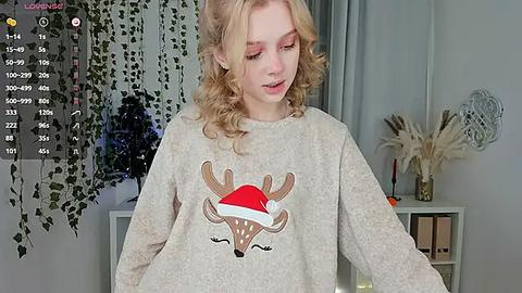 Media: A young woman with curly blonde hair, wearing a cozy gray sweatshirt featuring a red and white Santa hat with antlers, stands in a softly lit room with greenery and a white shelf in the background.