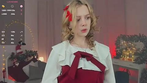 Media: Video of a young blonde woman in a festive red dress with a white ruffled collar, standing in a warmly lit room with Christmas decorations.