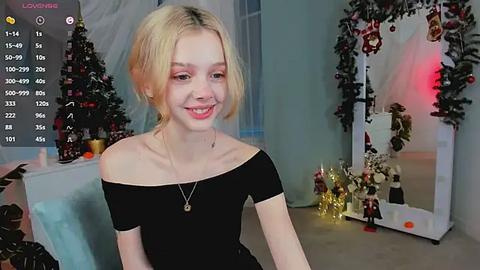 Media: Video of a smiling, blonde, fair-skinned young woman with an off-the-shoulder black top, standing in a festive room with Christmas decorations, including a lit tree and framed photos.