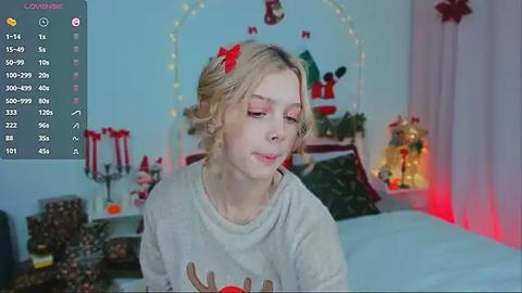 Media: Video of a young, fair-skinned woman with blonde hair, wearing a reindeer sweater, sitting in a cozy, festive bedroom with holiday decorations and a lit Christmas tree in the background.