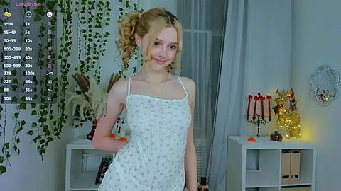 Media: Video of a young woman with curly blonde hair in a white floral dress, smiling in a cozy, softly lit room with hanging plants, candles, and a calendar.