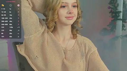 Media: Video of a young, fair-skinned woman with wavy, shoulder-length blonde hair, wearing a beige knit sweater, standing indoors with a blurred background.