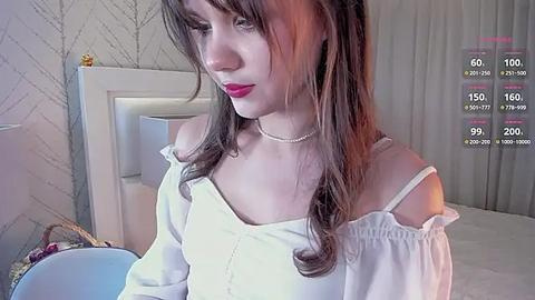 Media: Video of a young woman with long brown hair, wearing a white off-shoulder blouse and a pearl necklace, standing in a bedroom with light-colored walls and a white headboard.