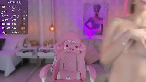 Media: Video of a pink gaming chair with a cartoonish face, set in a dimly lit room with purple accents, featuring a blurred person holding a phone.