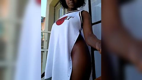 Media: Video of a dark-skinned woman with short curly hair, wearing a sleeveless white tank top with a red sun logo, exposing her bare chest and thighs. She stands outside on a balcony.