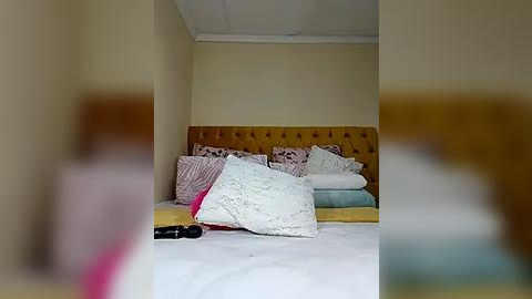 Media: Video of a cozy bedroom with a plush, tufted mustard-yellow headboard, neatly made bed with striped pillows, and a black vibrator on the white bedspread. The room features beige walls and a white ceiling.