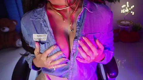 Media: Video of a woman with long dark hair, wearing a denim jacket and a pink thong, revealing ample cleavage, holding her breasts. Background includes a teddy bear and a glowing lamp.