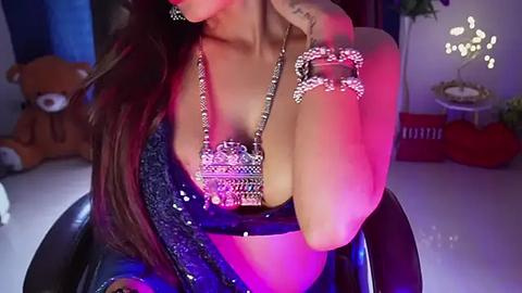 Media: Video of a woman with long brown hair, wearing a black sequined top and a large, ornate silver necklace, sitting in a dimly lit room with purple lighting, plush toys, and a decorative tree in the background.