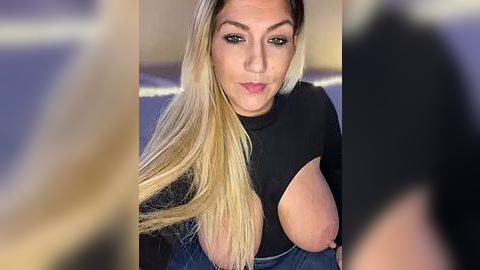 Media: A video of a blonde woman with long, straight hair, wearing a black top exposing one large breast, taken indoors with a blurred background.