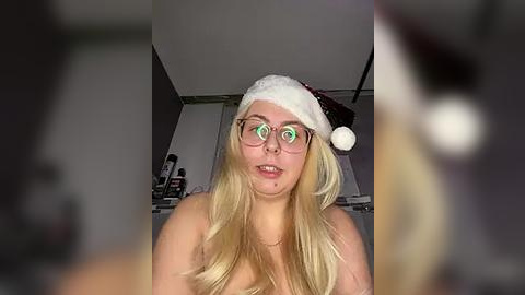 Media: Video of a young woman with long blonde hair, wearing a Santa hat, glasses, and a necklace, smiling in a dimly lit room.