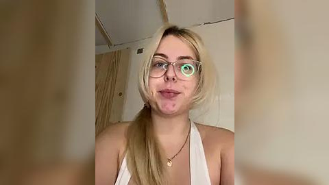 Media: Video of a young, fair-skinned woman with blonde hair in a ponytail, wearing glasses, a white halter top, and a heart pendant. She has a playful, slightly puckered face.