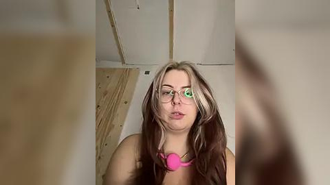 Media: Video of a fair-skinned, long-haired young woman with glasses and a pink necklace, standing in a room with wooden beams and unfinished walls.