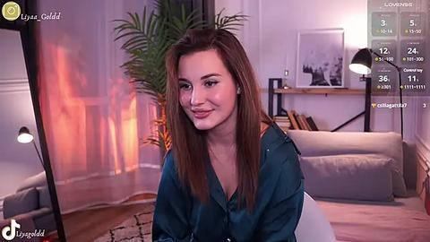 Media: Video of a smiling, fair-skinned woman with long, straight brown hair, wearing a teal blouse, seated in a cozy living room with a potted plant, wooden floor, and modern decor.