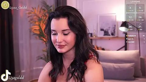 Media: Video of a woman with long black hair and fair skin, wearing a red top, smiling softly. Background shows a modern living room with a plant and a clock.