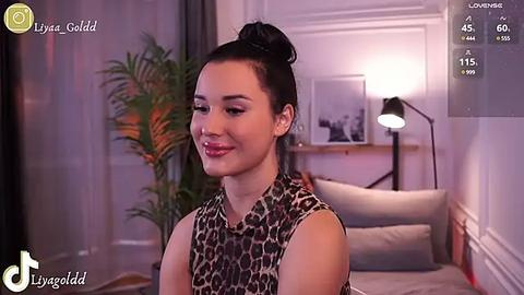 Media: Video of a smiling woman with fair skin, dark hair in a bun, wearing a leopard print top, in a modern bedroom with a potted plant, lamp, and framed picture.