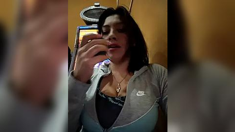 Media: Video of a young woman with dark hair, wearing a light blue Nike jacket and black top, taking a selfie with an old flip phone. Background is blurred.