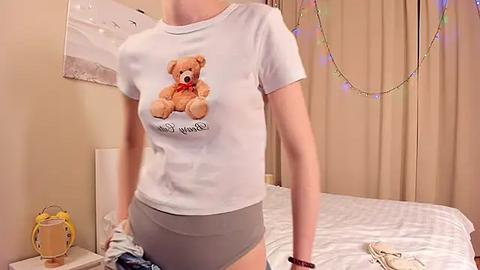 Media: Video of a fair-skinned woman wearing a white T-shirt with a teddy bear graphic and grey panties in a cozy bedroom with white sheets, beige curtains, and string lights.