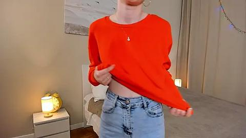 Media: Video of a slender, fair-skinned person in a bright red long-sleeve shirt, blue jeans, lifting the shirt to reveal their stomach. Background features a beige wall, a lamp, and a beige chair.