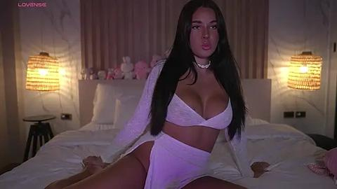 Media: Video of a dark-haired woman in white lingerie, sitting on a bed with white sheets, in a dimly-lit, elegant bedroom.