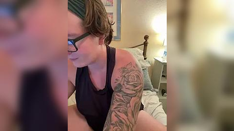 Media: Video of a fair-skinned woman with short, wavy brown hair, glasses, and a black tank top, sporting a colorful tattoo sleeve, sitting in a softly lit bedroom.