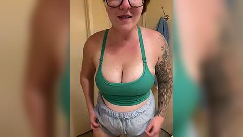 Media: Video of a plus-sized woman with glasses, wearing a green sports bra and grey sweatpants, showcasing a large tattoo sleeve on her left arm. The background features a beige wall and a blue towel.