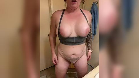 Media: Video of a plus-sized woman with fair skin and medium-sized breasts in a bathroom. She wears a gray sports bra and beige panties. Her left arm has a tattoo sleeve. Background includes beige walls, towels, and a shower curtain.