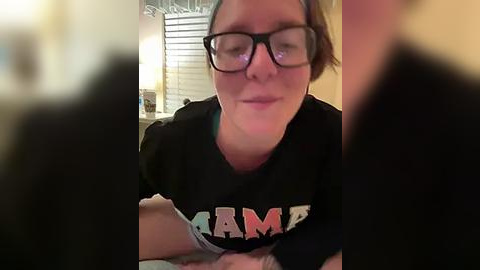 Media: A candid video of a woman with glasses and short brown hair, wearing a black \"AMAZING\" t-shirt, sitting indoors, slightly out of focus.