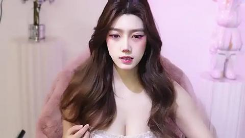 Media: Video of a young East Asian woman with long, wavy brown hair, fair skin, and pink eyeshadow. She wears a lace bralette, a fur shawl, and has a confident expression. Background features floral decor and soft pink lighting.