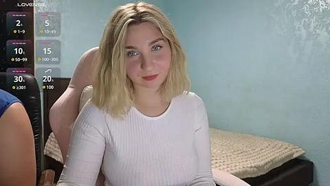 Media: Video of a young woman with blonde hair, wearing a white ribbed top, sitting on a bed in a dimly lit room.