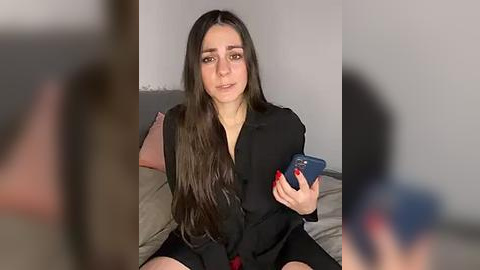 Media: Video of a young woman with long, dark hair, wearing a black blazer and red shorts, holding a smartphone, sitting on a bed with a grey pillow and beige sheets, blurry background.