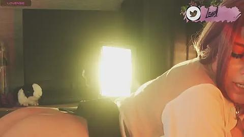 Media: A video of a woman with shoulder-length brown hair, wearing a white top, lying on her side in a dimly-lit room with a bright, glowing object in the background.