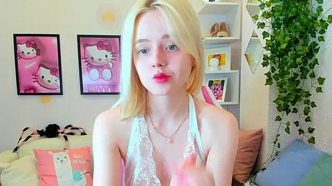 Media: Video of a blonde young woman with fair skin and blue eyes, wearing a white lace bra, posing in a pink-themed bedroom with Hello Kitty decor and green ivy.