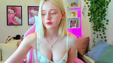 Media: Video of a fair-skinned, blonde woman with straight hair, wearing a white lace bra, seated on a pink gaming chair, surrounded by Hello Kitty decor, in a cozy room with green plants and neon lights.